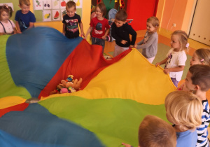 dzieci na dywanie stoją z kolorową chustą animacyjną trzymają jej brzegi, na niej leża małe maskotki
